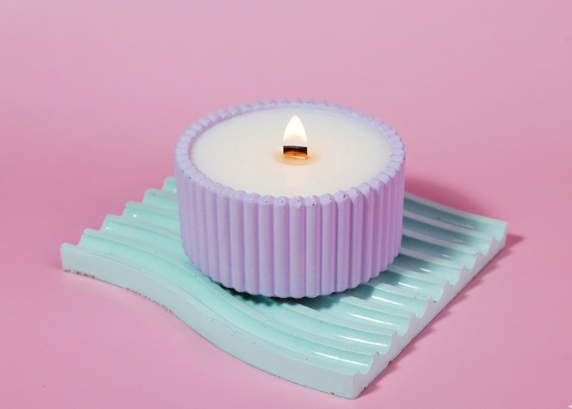 A lit wick candle in a ribbed purple handmade lavender purple candle holder, resting on a mint green wavy candle tray against a solid pink background. The candle is made with soy wax and a wooden wick and has eucalyptus lavender cedar and rose
