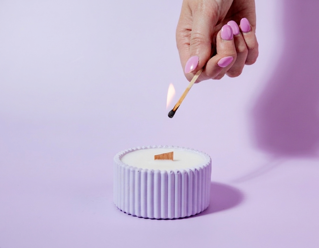 A hand with pink nail polish holds a lit match above a small purple candle in a ribbed light purple container against a lavender background.