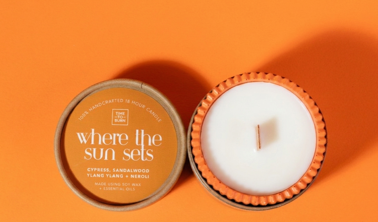 An orange jar candle and its lid are arranged on a solid orange background. The lid is tan with the text 'where the sun sets' and 'orange, cYPRESS, SANDALWOOD + NEROLI'.