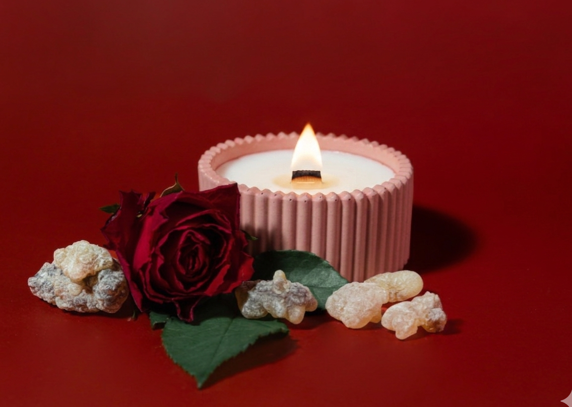 A pink ribbed candle with a lit flame sits on a red background, styled with a dark red rose, green leaves, and small beige stones.
