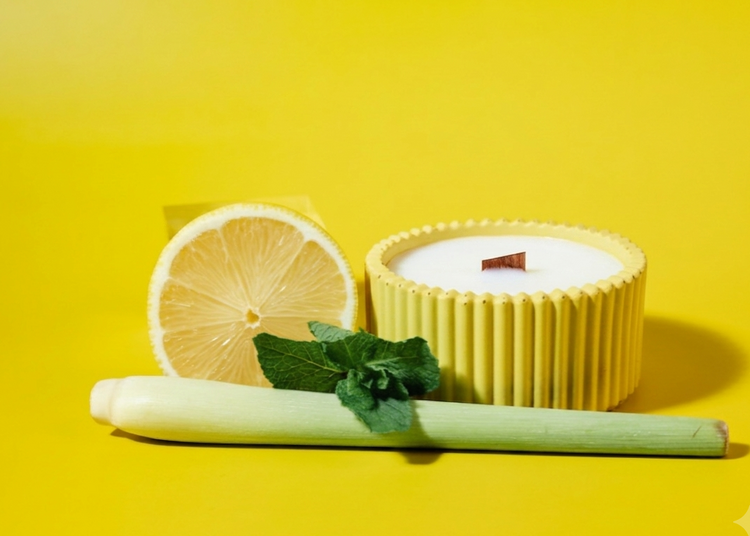 A yellow citronella ribbed handmade candle jar with white soy wax sits on a bright yellow background, styled with a sliced lemon, a green stalk, and mint leaves.