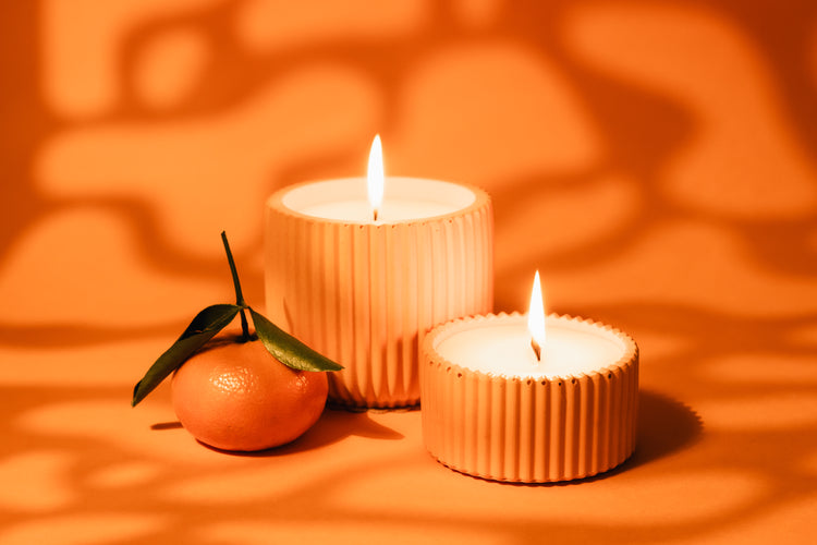 A hand with pink nail polish holds a lit match above a small purple candle in a ribbed light purple container against a lavender background.