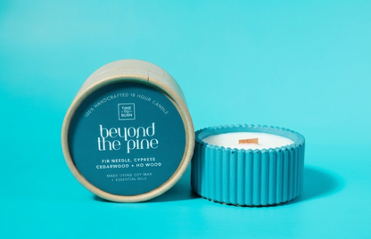 A blue scented candle by time to burn named 'Beyond the Pine', shown with its lid off to reveal the white wax and wick. The candle is set against a solid bright turquoise-blue background.