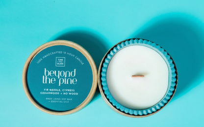 An open teal and beige candle tin on a bright blue background, showing white soy wax inside and the lid label reading 'beyond the pine' and 'Tine Tree Candle'.