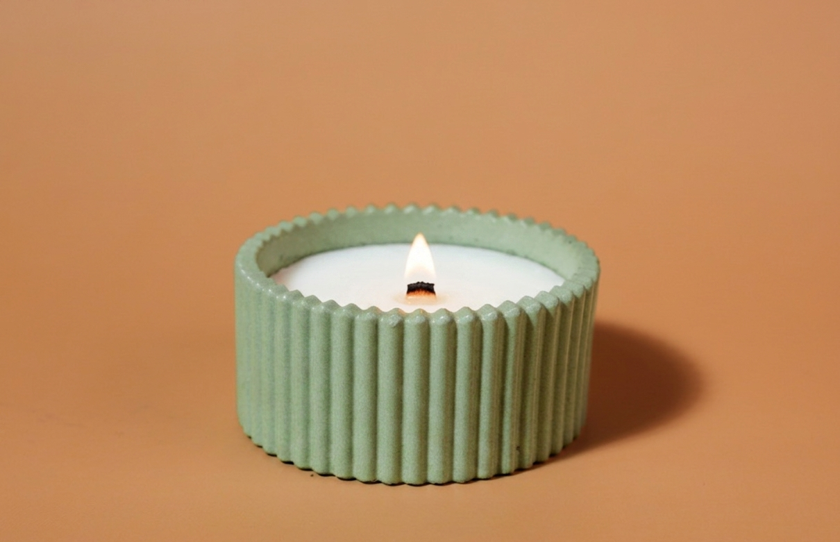 A lit handmade candle in a ribbed sage green holder, set against a solid warm tan background. Vetiver and thyme