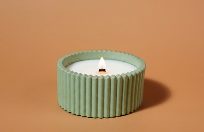 A lit handmade candle in a ribbed sage green holder, set against a solid warm tan background. Vetiver and thyme