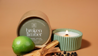 A candle product styled on a warm tan background with a lime, wood pieces, and peppercorns. The set includes a kraft paper-wrapped jar labeled 'broken timber' and a mint green ribbed candle holder with a lit candle.