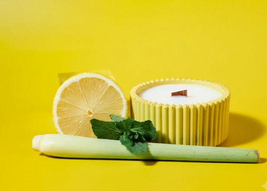 A yellow citronella ribbed handmade candle jar with white soy wax sits on a bright yellow background, styled with a sliced lemon, a green stalk, and mint leaves.