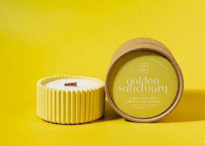A candle product shown on a bright yellow background, with one ribbed yellow candle container open to reveal white wax and wick, and another cylindrical container standing beside it with a round lid label reading 'golden sanctuary' and 'citronella, mint + palmarosa'.