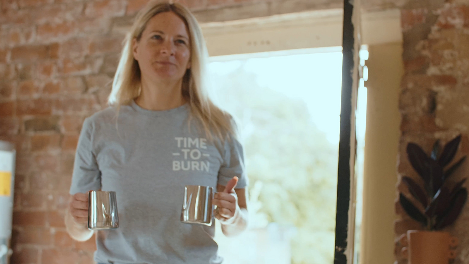 A woman with blonde hair stands in a doorway holding two metallic mugs, wearing a gray t-shirt with the text 'TIME TO BURN' printed on the front. The background features a rustic brick wall and a bright, sunlit interior.