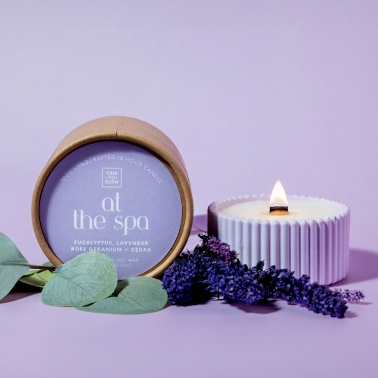A spa-themed candle set on a lavender background, featuring a round wooden-rimmed tin labeled 'at the spa' and a white ribbed candle with a lit flame, styled with green leaves and purple lavender sprigs.