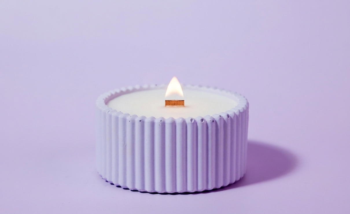 A small purple lavender candle in a ribbed, cup-like holder with a lit wooden wick, centered on a solid light purple background.