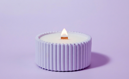 A small purple lavender candle in a ribbed, cup-like holder with a lit wooden wick, centered on a solid light purple background.