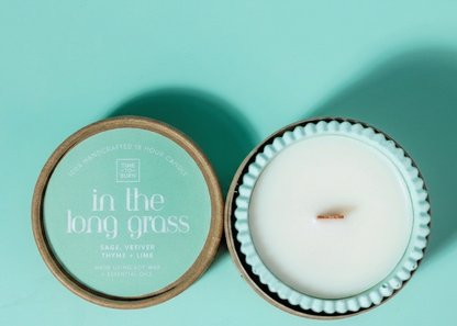 A green wooden wick scented candle in a light green handmade candle jar next to its round lid, which features a mint green label reading 'in the long grass'. Candle contains scents of vetiver thyme sage and thyme 