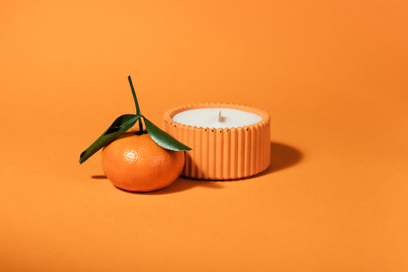 A small orange-shaped candle in a ribbed terracotta holder sits beside a real orange with green leaves on a solid orange background.