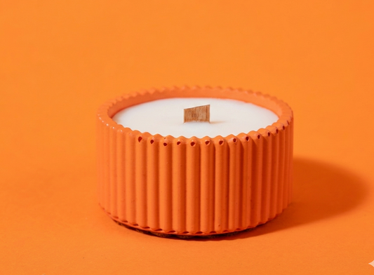 A small candle in a ribbed orange container with a white wax surface and a single wooden wick, centered on a solid orange background.