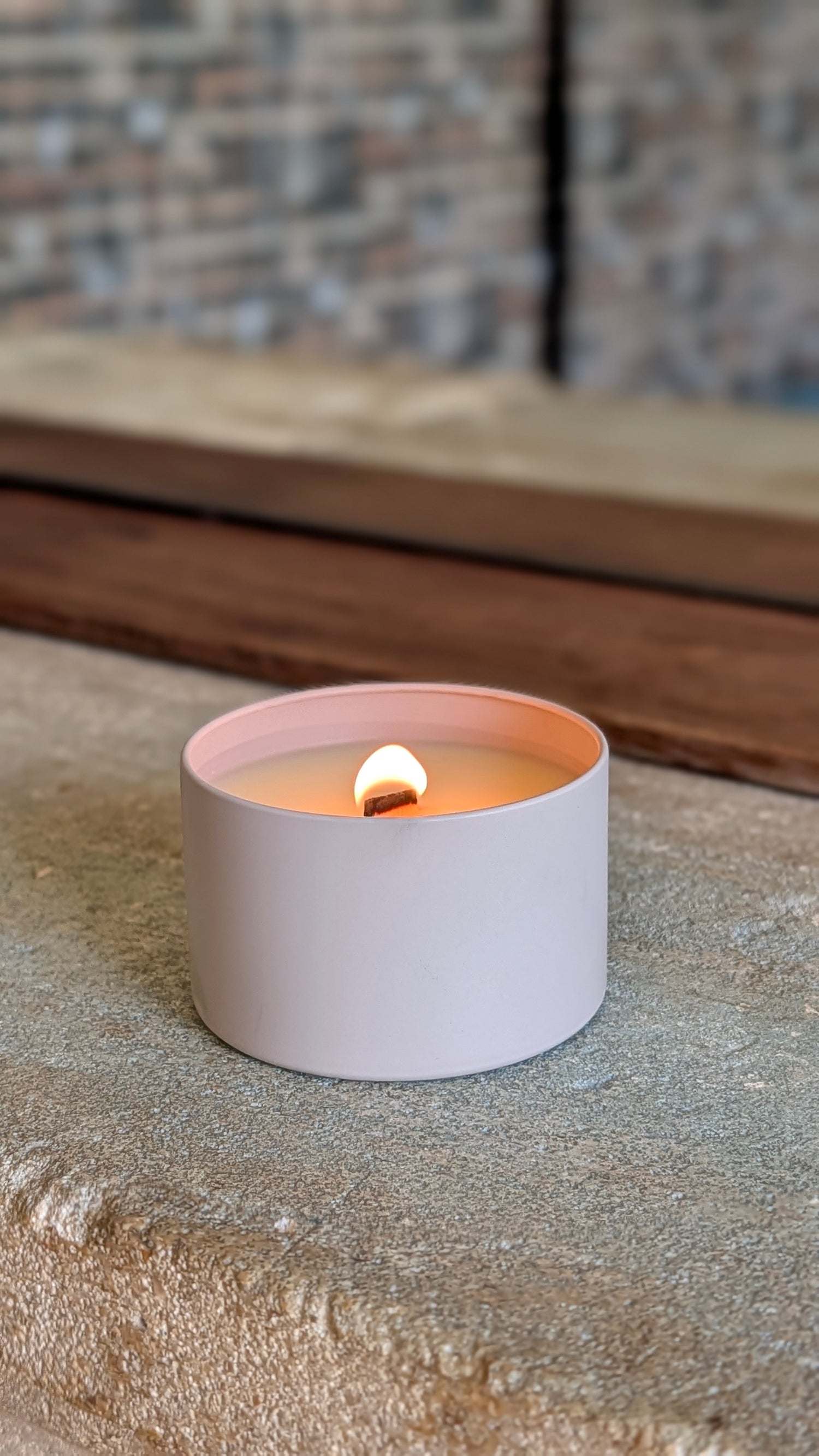 A minimalist matte pale pink candle in a cylindrical tin container, lit with a single flame, resting on a textured stone surface with a rustic wooden and brick background.
