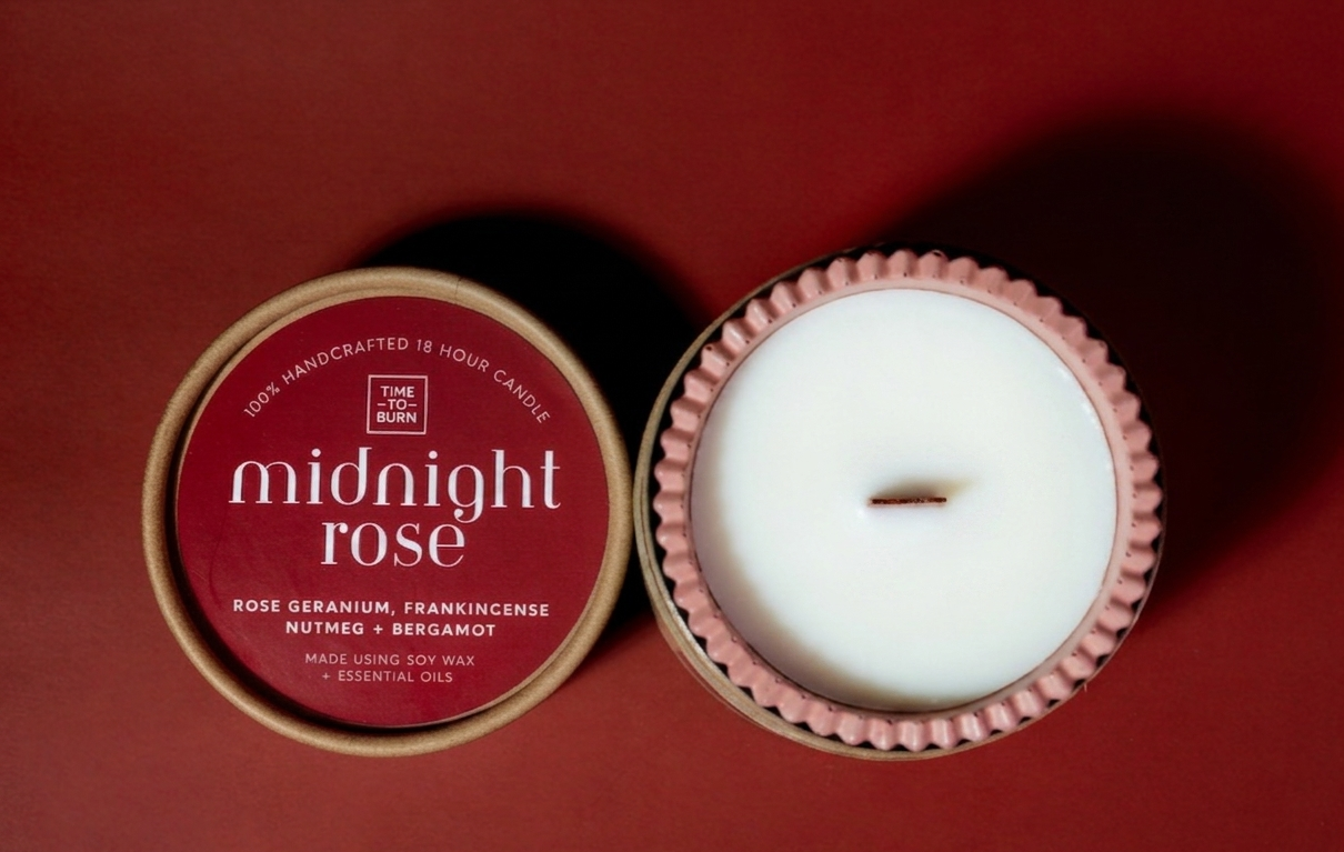 An open scented candle in a red scalloped handmade candle jar with white soy wax, shown beside its lid on a dark red background. The lid label reads 'midnight rose' and includes Frankinsense and rose geranium.