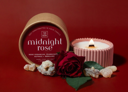 A candle product display on a dark red background, featuring a kraft paper can labeled 'midnight rose' next to a lit pink ribbed candle holder. A red rose with green leaves and small white stones are arranged in front.