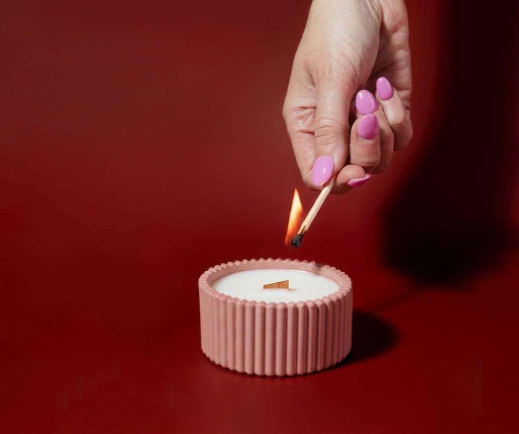A hand with pink nail polish holds a lit match above a small ribbed pink candle holder containing white wax, set against a solid dark red background.