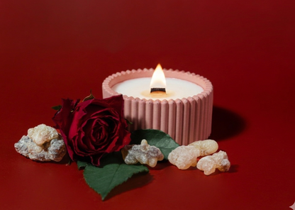 A pink ribbed candle with a lit flame sits on a red background, styled with a dark red rose, green leaves, and small beige stones.
