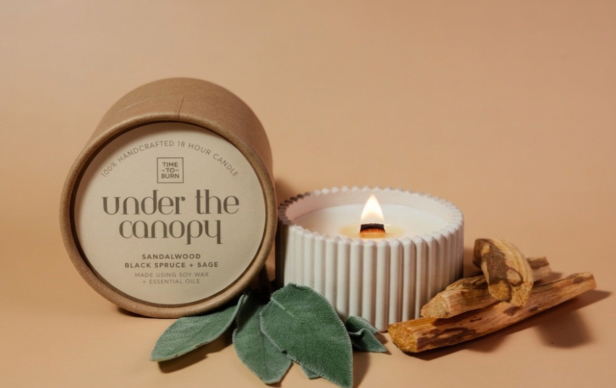 A candle product styled on a warm beige background, showing a kraft paper tub labeled 'under the canopy' next to a white ribbed candle holder with a lit wick, plus sage leaves and dried wood pieces.