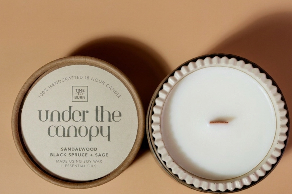 A beige simple coloured candle with handmade jar shown next to its lid on a warm beige background. The candle wax is white.and soy, and the label reads 'under the canopy' with 'SANDALWOOD BLACK SPRUCE + SAGE' below it.