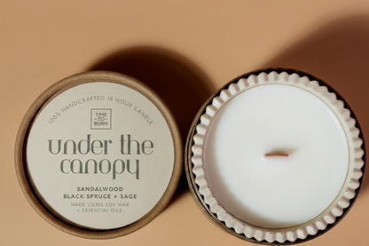 A beige simple coloured candle with handmade jar shown next to its lid on a warm beige background. The candle wax is white.and soy, and the label reads 'under the canopy' with 'SANDALWOOD BLACK SPRUCE + SAGE' below it.